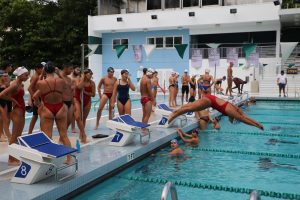 Several members of the Cavalier Swimming team stand beside Ransom Everglades High Schools pool as others plunge into the water.