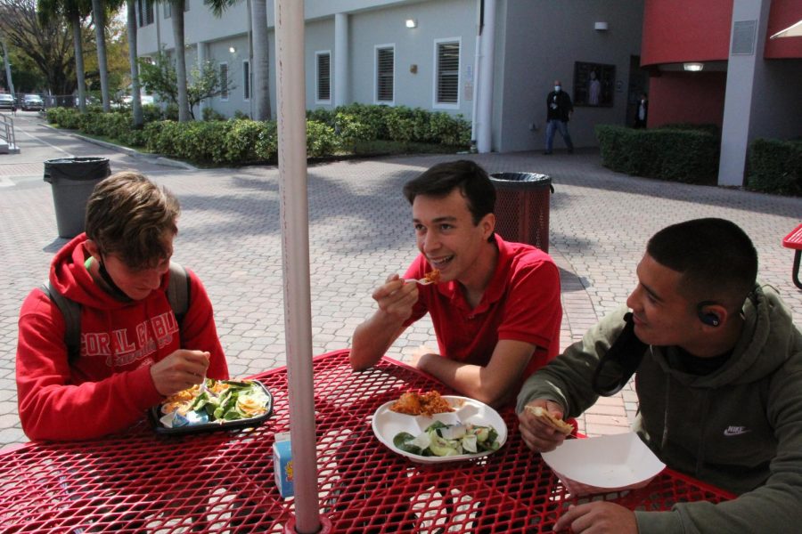 A group of sophomore boys converse while enjoying the school lunch. Their plates are filled with chicken tenders, spaghetti and cheese sticks. 