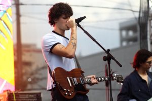 A School of Rock student performs Latin music at Carnaval Miami 2025 with both a guitar and microphone in hand.